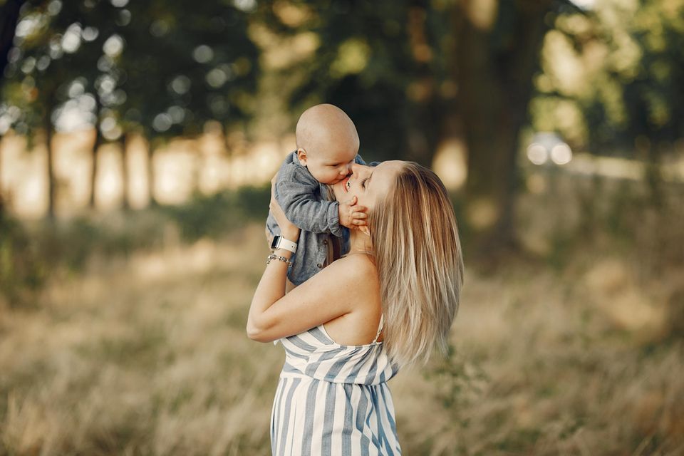 Eine blonde Frau in einem gestreiften Kleid hält ein Baby in den Armen und befindet sich in der Natur