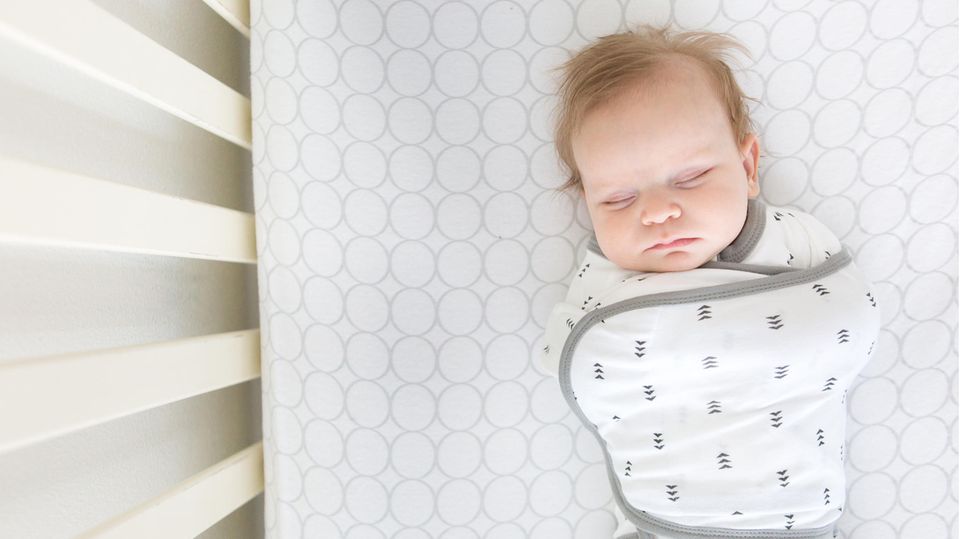 Ein Baby liegt in einem Babyschlafsack mit geschlossenen Augen im Bett.