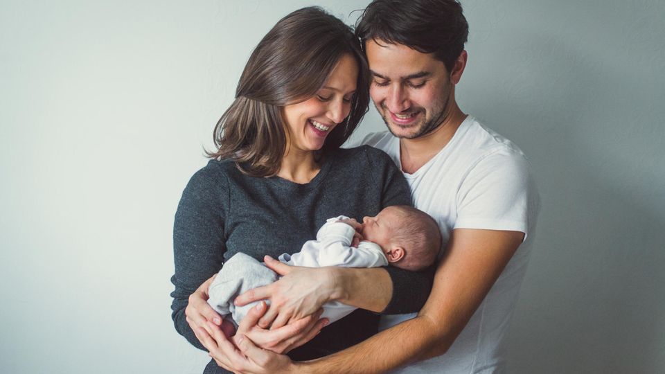 ein junges Elternpaar hält ein Neugeborenes in den Armen und lächelt es an.