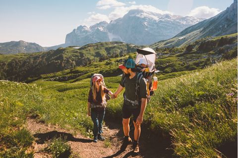 Kraxe im Test: Junges Paar wandert mit Baby in der Kraxe in sommerlicher Alpenlandschaft.