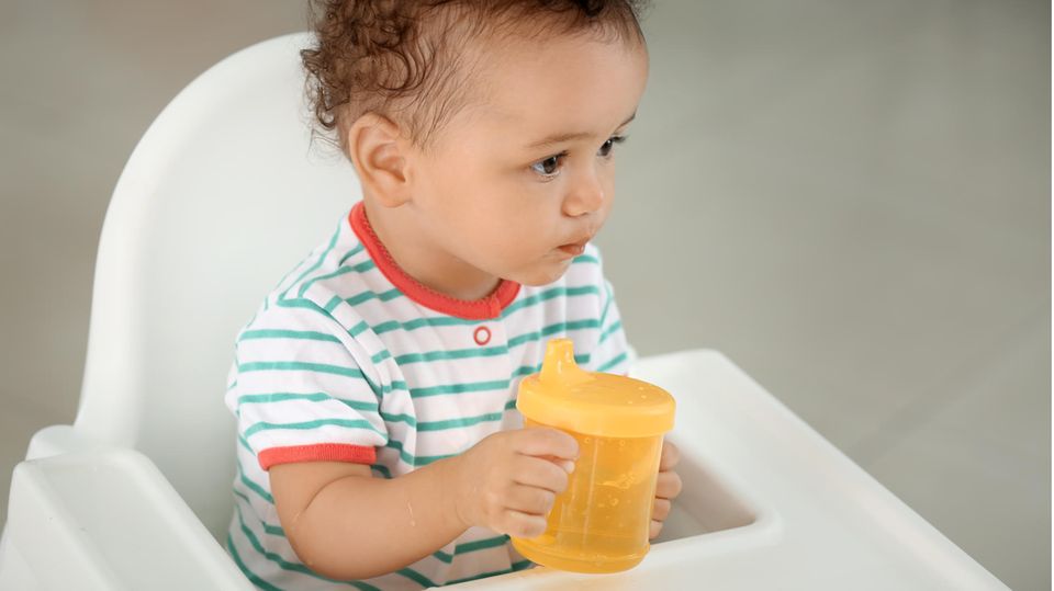 Ein Kleinkind sitzt in einem Babystuhl und hält einen gelben Trinkbecher in den Händen