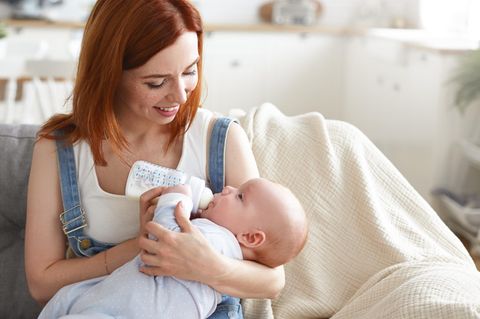 Trinkmenge Baby: Eine Mutter hält ihr Baby auf dem Arm und gibt lächeln das Fläschchen