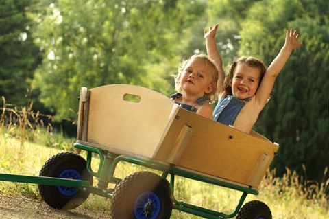Zwei Kleinkinder sitzen fröhlich in einem Holz-Bollerwagen bei Sonnenschein in der Natur.