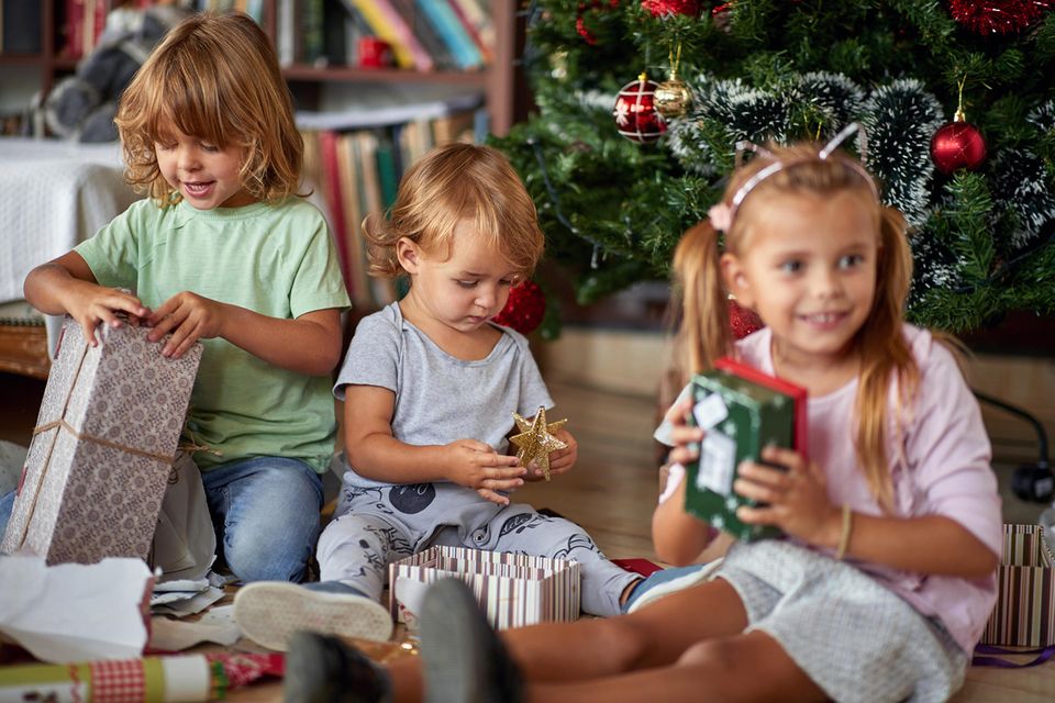 Weihnachtsgeschenke für Kinder: Drei kleine Kinder packen unter dem Weihnachtsbaum Geschenke aus.