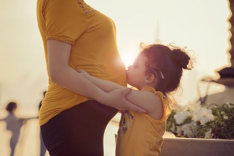 Kleines Mädchen küsst im Sonnenlicht den schwangeren Bauch ihrer Mutter