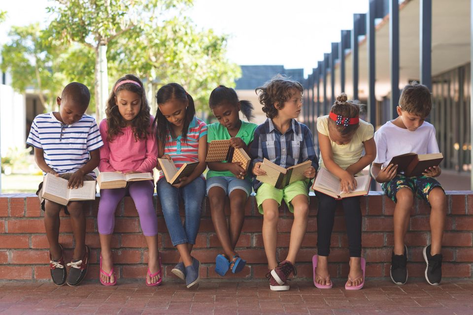 Kinderbücher über Geflüchtete: Verschiedene Kinder sitzen zusammen auf Steinmauer und lesen.