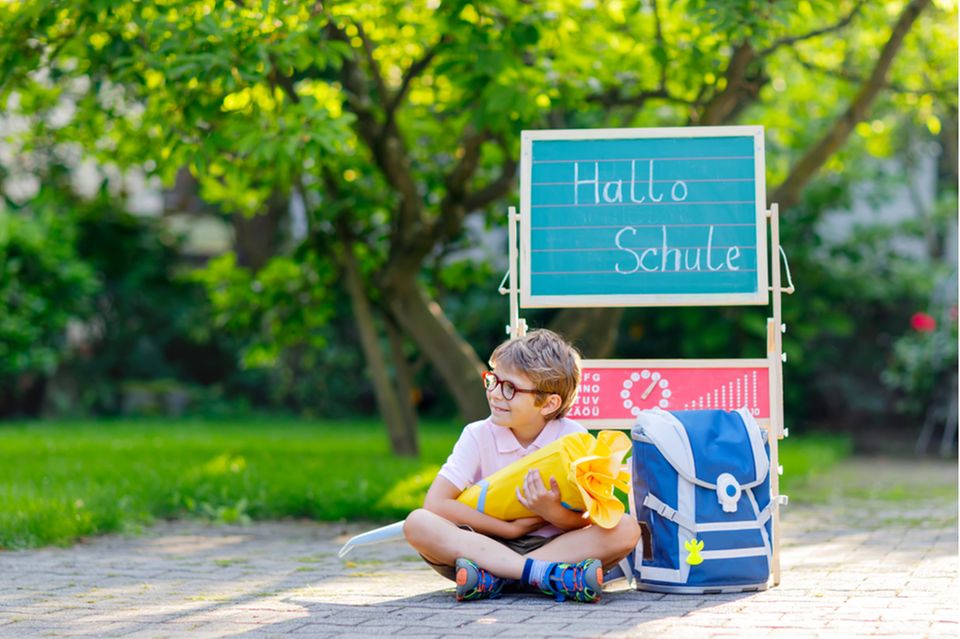 Einschulung: Junge mit Brille sitzt mit Schultüte und Schulranzen auf dem Schulhof.