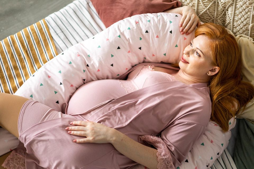 Hochschwangere Frau liegt verträumt mit einem Stillkissen im Bett