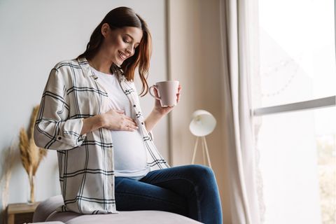 Schwangerschaftsdauer: Eine schwangere Frau sitzt mit einer Tasse Tee lächelnd auf dem Sofa und berührt ihren Bauch.
